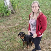 Julija steht auf einer Wiese neben einem sitzenden Rottweiler, sie trägt eine rote Jacke, ein helles T-Shirt und dunkle Hosen.