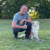 Wolfgang hockt auf einer Wiese neben einem sitzenden Hund mit hellem Fell, beide blicken nach vorne, vor ihnen liegen zwei Leinen auf dem Gras.