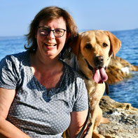Silke sitzt am Felsen am Meer, trägt ein kurzärmeliges, gemustertes Shirt, neben ihr sitzt ein gelber Labrador mit heraushängender Zunge.
