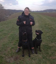 Claudi steht auf einem grasbewachsenen Weg, trägt schwarze Outdoorjacke und Hose, zwei schwarze Hunde sitzen vor ihr und schauen zu ihr hoch.