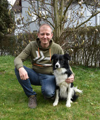 Andi Baumann kniet auf Gras und hält seinen schwarz-weißen Border Collie namens Balu an der Seite.