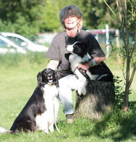 Sigi sitzt auf einem Baumstumpf im Gras, hält eine schwarz-weiße Border-Collie-Hündin auf dem Schoß, ein weiterer Border-Collie sitzt neben ihm auf dem Boden.