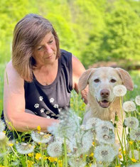 Andrea sitzt auf einer Wiese mit Löwenzahn und hält ihre Hündin Sunny, die nach rechts schaut.