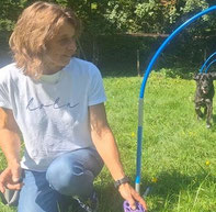 Lisa kniet auf einer Wiese, trägt ein weißes T-Shirt mit dem Schriftzug 'leben' und hält ein lila Hundespielzeug, im Hintergrund läuft ein schwarzer Hund durch einen blauen Hoopers-Reifen.
