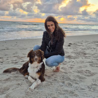 Hüda in schwarzer Jacke und blauer Jeans kniet am Strand neben braun-weißem Hund, im Hintergrund Meer und bewölkter Sonnenuntergang.