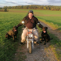 Josip sitzt auf einem kleinen Motorrad auf einem Feldweg, neben ihm zwei schwarze Hunde, umgeben von grünen Wiesen und Bäumen im Hintergrund.