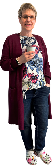 Silke Uden in Jeans, buntem Shirt und dunkelroter Strickjacke hält eine Kaffeetasse in der Hand und lächelt in die Kamera.