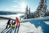 Vier Personen auf Skiern stehen am Pistenrand und schauen auf ein verschneites Bergpanorama über einem Meer aus Wolken.
