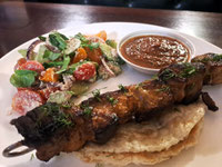 White plate with skewer of pork, bowl of red sauce and mixed vegetable salad