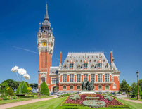 Vue extérieure de l'Hôtel de Ville de Calais et de son beffroi (patrimoine UNESCO), avec le jardin fleuri et la statue des Bourgeois de Calais au premier plan, sous un ciel bleu ensoleillé.
