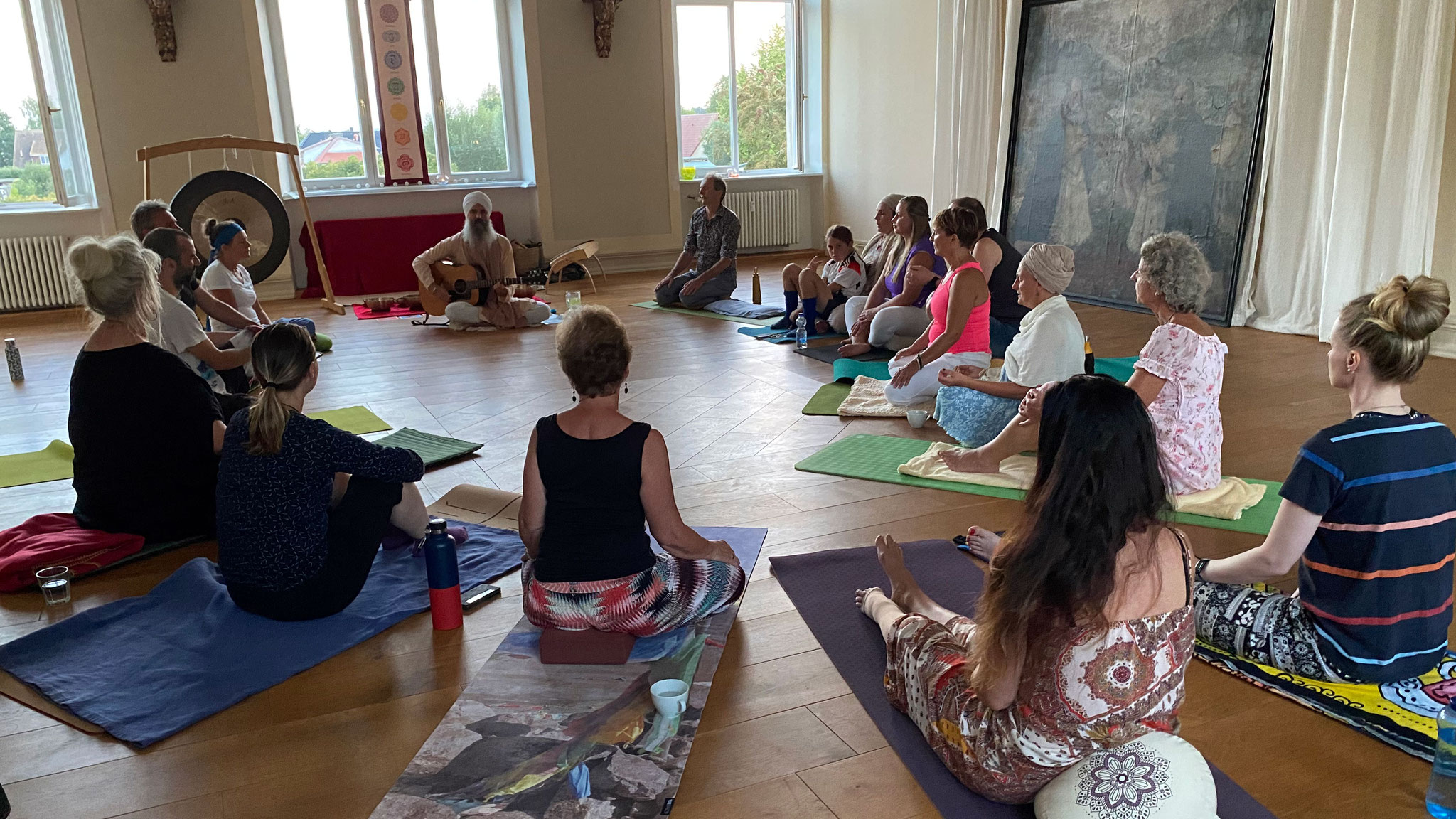 Mehrere Frauen und Männer sitzen im Kreis in meditativer Haltung, ein Mann mit Turban spielt Gitarre