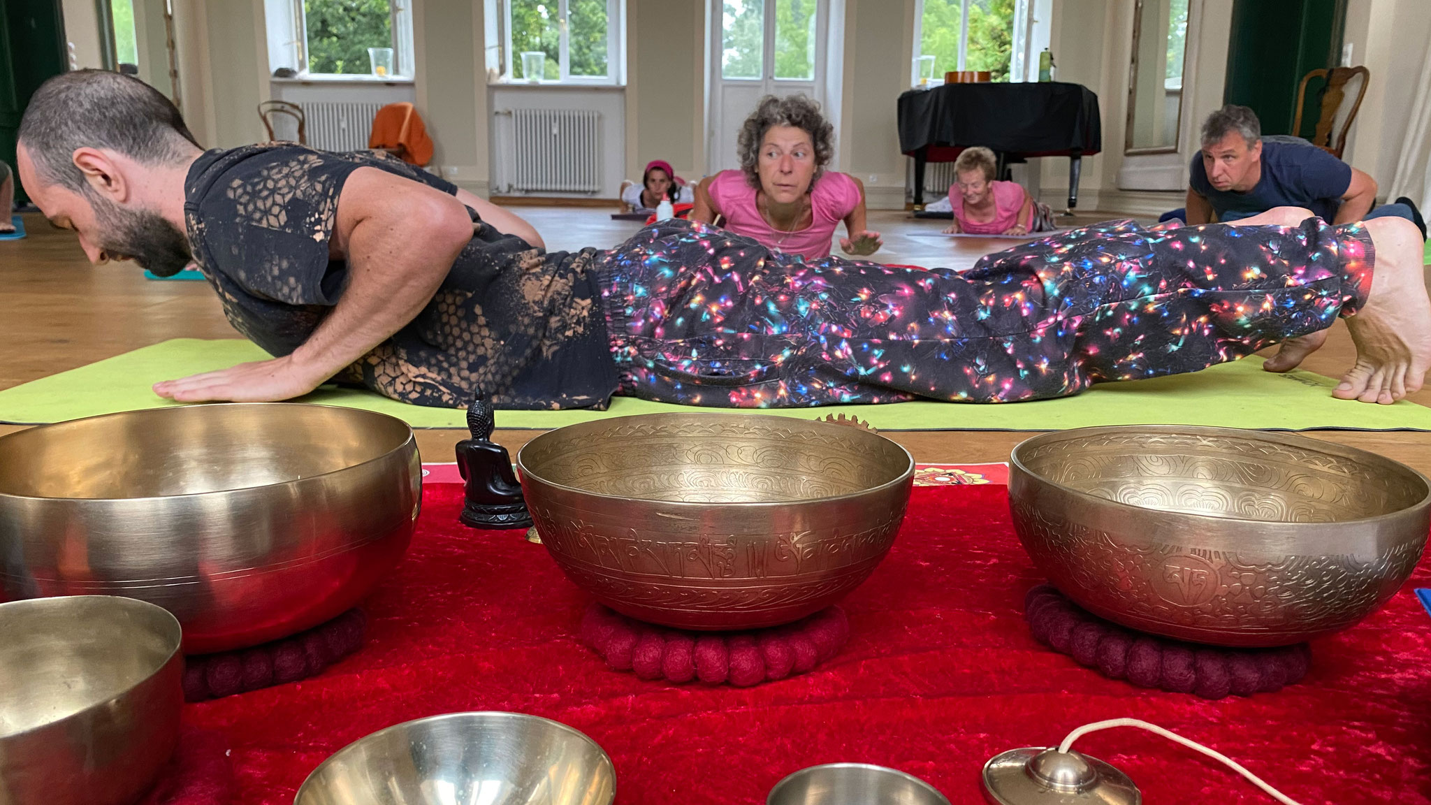 Im Vordergrund Klangschalen auf rotem Teppich, dahinter Seminarteilnehmer in einer liegenden Yogaposition  