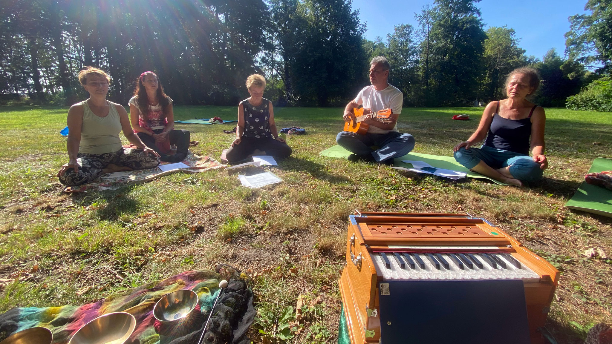 Sitzende Menschen auf einer sommerlichen Wiese in meditativer Haltung, im Vordergrund ein Tasteninstrument 