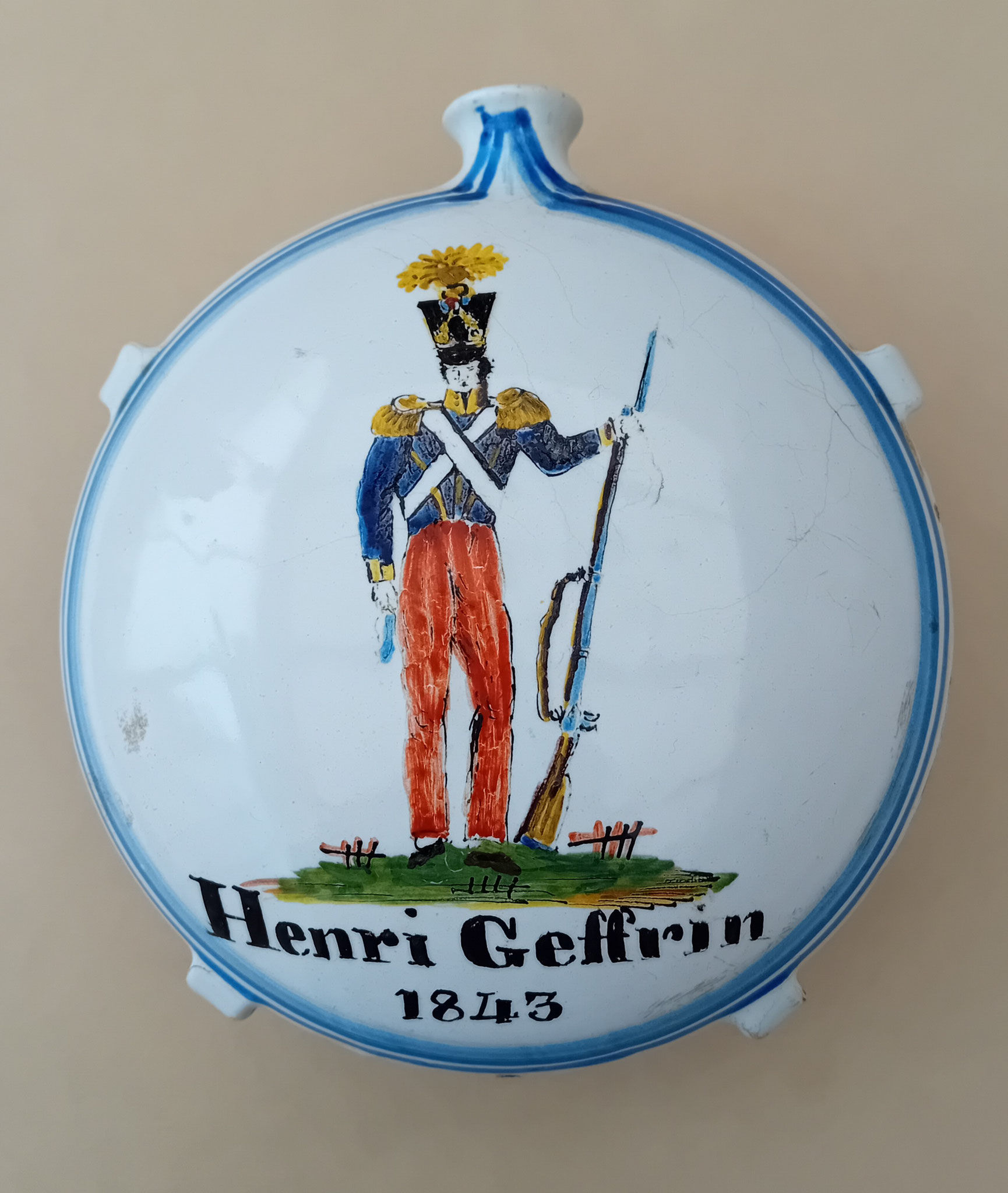 Gourde Diam. 18 : cette gourde crapaud patronymique "Henri Geffrin 1843" laisse presque toute la place à un soldat en uniforme et fusil au pied, sur un tertre.