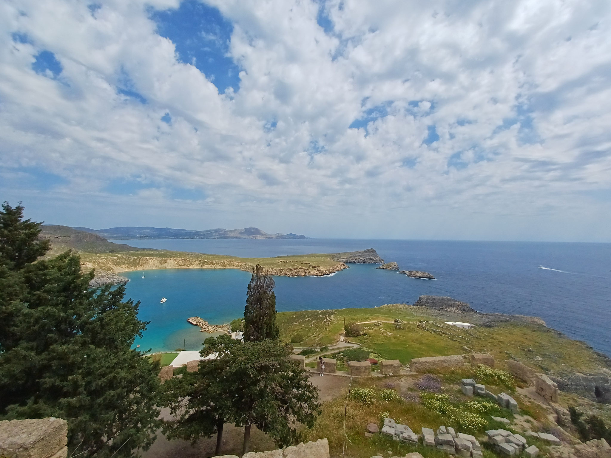 Ausblick über das türkisblaue Mer und einen Strand auf der linken Seite. Im Vordergrund sieht man Reste der Akropolis in einem Feld.
