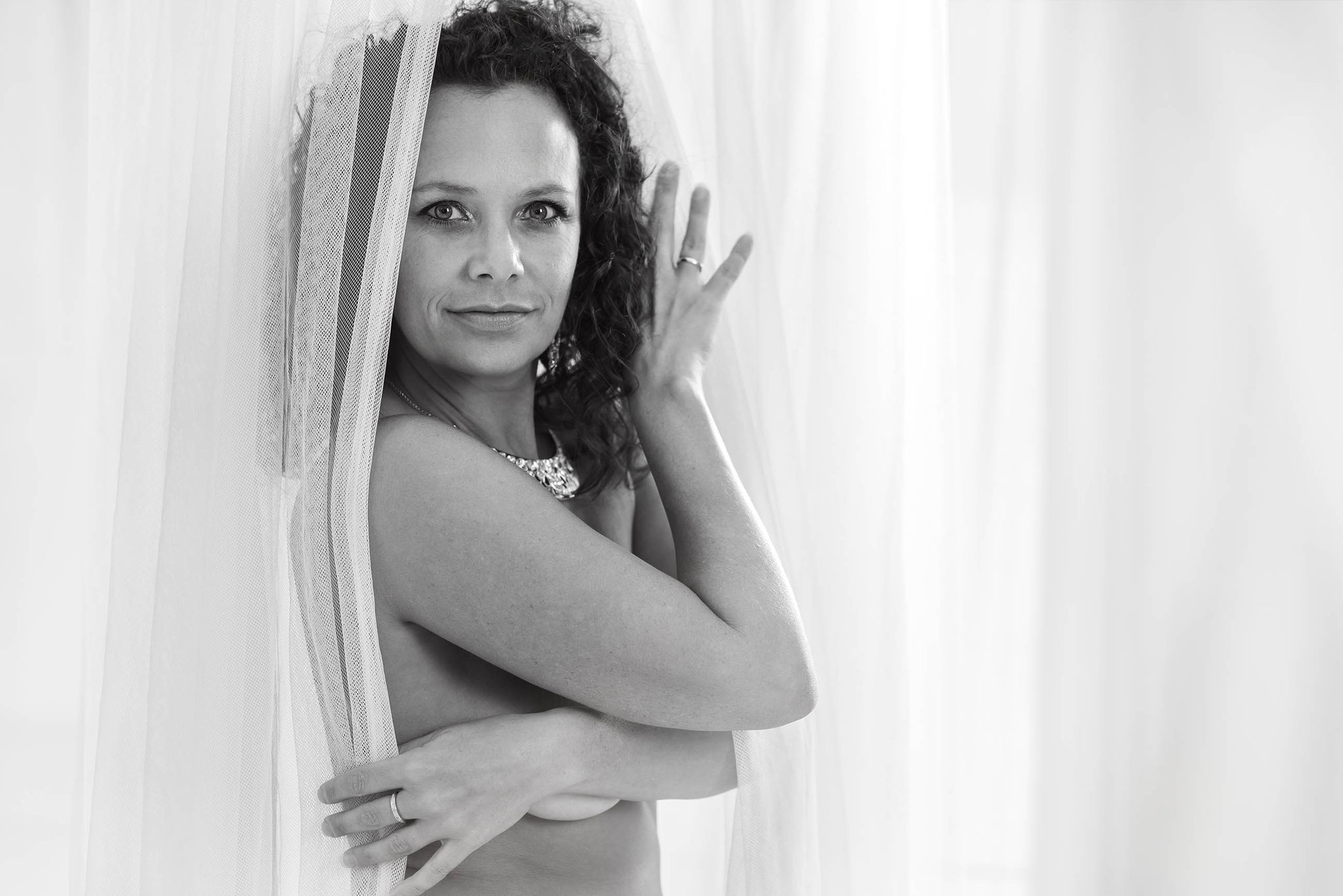 Eine selbstbewusste Frau mit lockigen dunklen Haaren blickt sanft in die Kamera, elegant mit einem weißen Vorhang bedeckt – sinnliches schwarz-weiß Portrait“