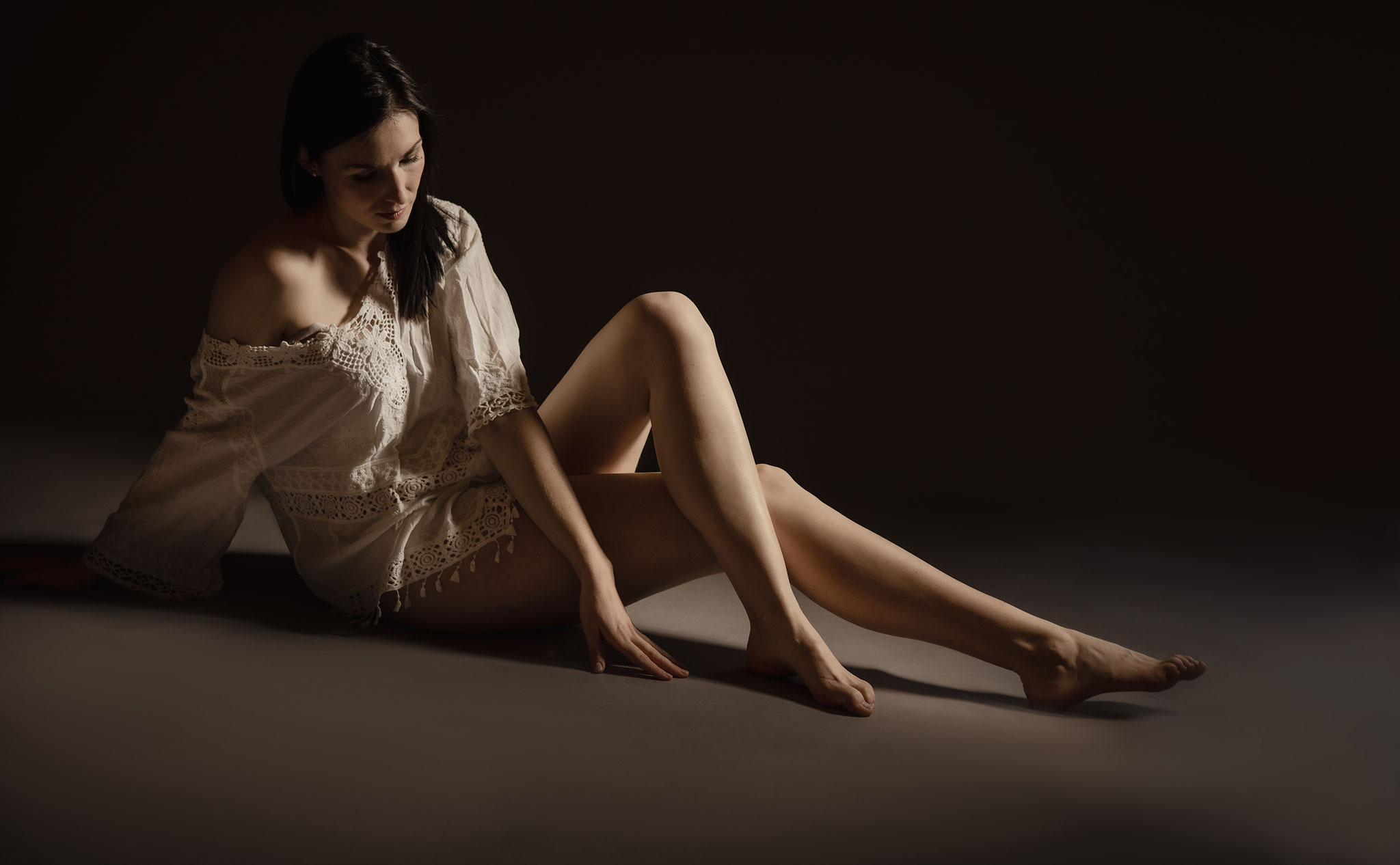 Sinnliches Studiofoto einer sitzenden Frau in heller Spitzenbluse, die im warmen Low-Key-Licht nachdenklich zu Boden blickt