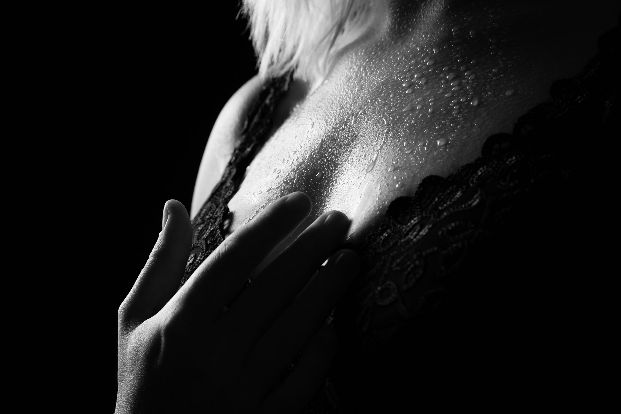 Detailreiches Schwarz-Weiß-Boudoirfoto eines weiblichen Dekolletés in Spitzenwäsche, mit Wassertropfen auf der Haut und sanft aufgelegter Hand