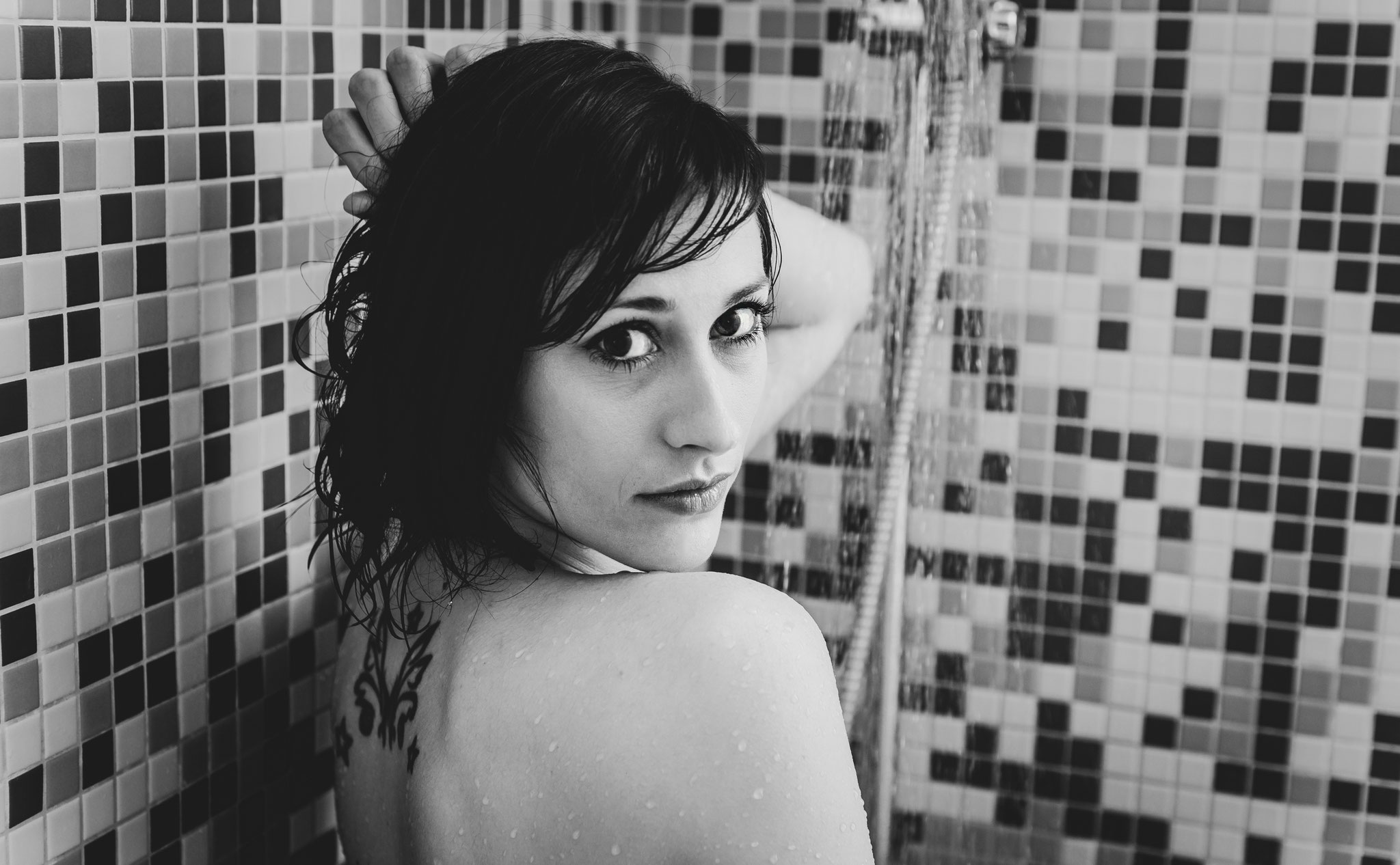 Sinnliches Schwarz-Weiß-Homeshootingfoto einer nackten Frau mit nassen Haaren unter der Dusche, die über die Schulter in die Kamera blickt