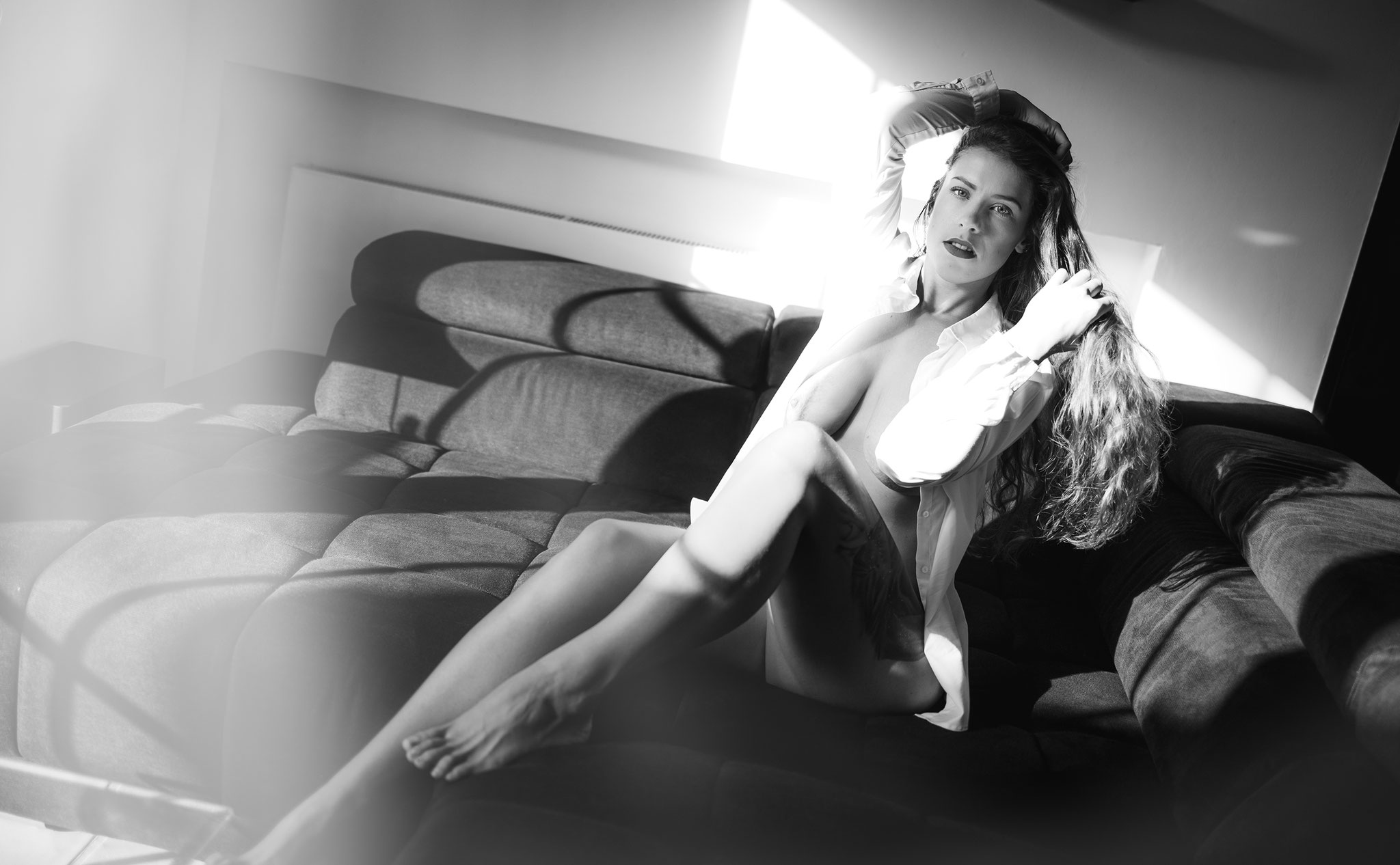 Sinnliches Schwarz-Weiß-Homeshootingfoto einer Frau in offenem weißen Hemd, die auf einem Sofa im dramatischen Spiel von Licht und Schatten posiert