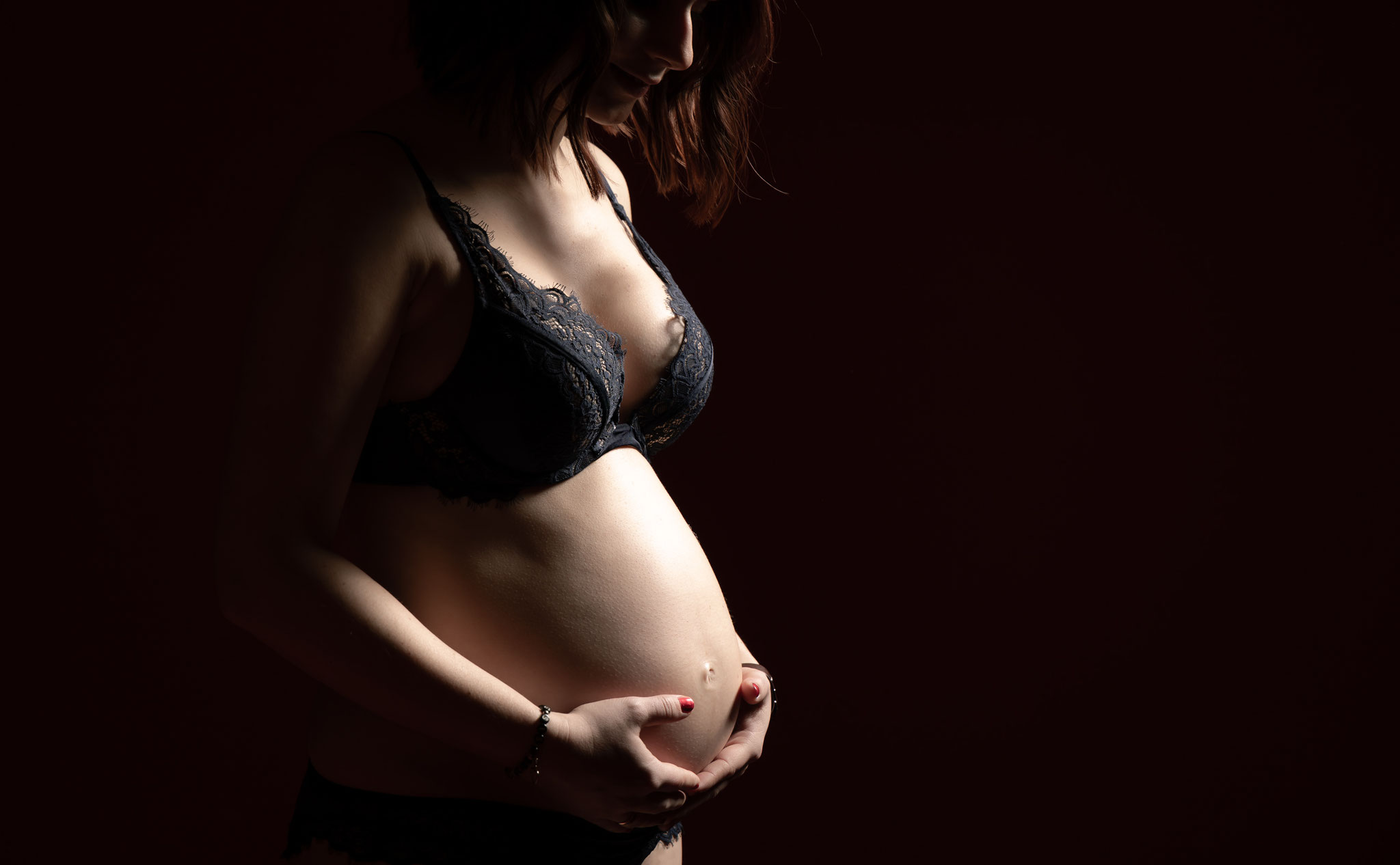 Stimmungsvolles Boudoir-Mutterschaftsfoto einer schwangeren Frau in schwarzer Spitzenunterwäsche, die im dunklen Studiolicht ihren Babybauch sanft mit beiden Händen umfasst