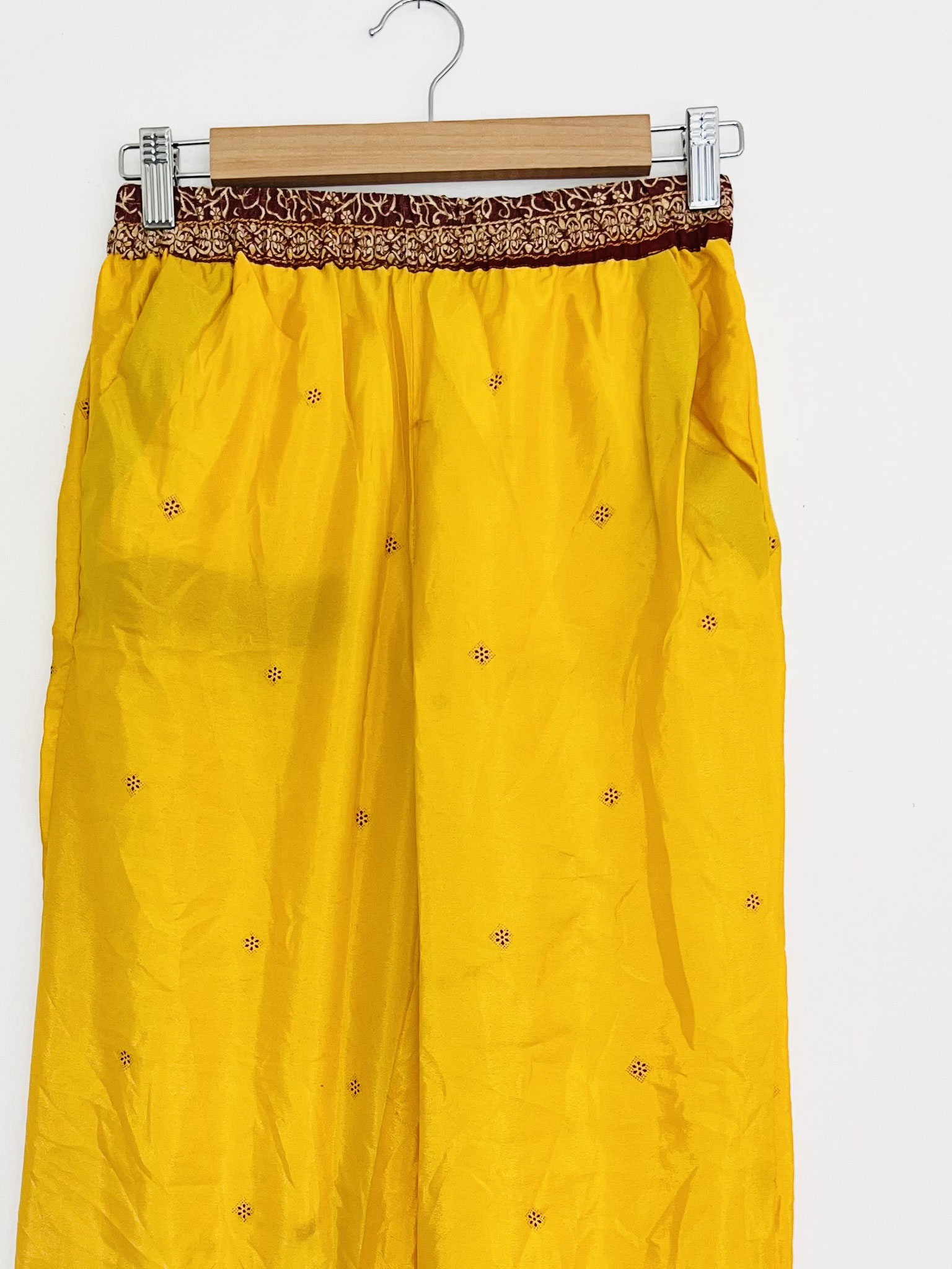 Vu de près sur cintre, ce pantalon met en valeur la richesse de son textile issu de saris recyclés. Les motifs bordeaux contrastent harmonieusement avec la teinte jaune moutarde et soulignent le caractère unique de cette pièce artisanale. Chaque détail ré