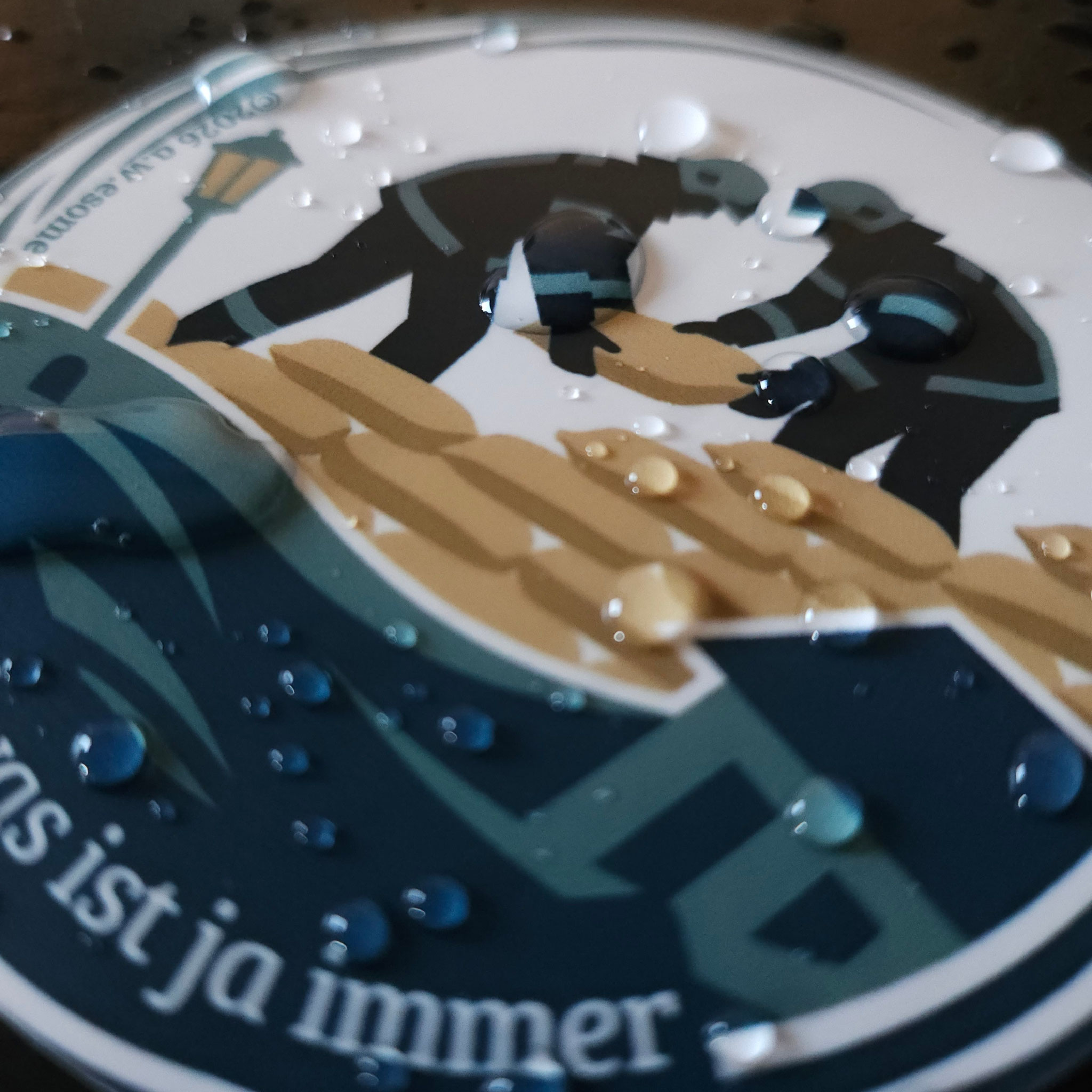 Wassertropfen auf rundem Feuerwehr-Sticker zum Sammeln. Motiv zeigt Einsatzkräfte mit Sandsäcken beim Hochwasserschutz.