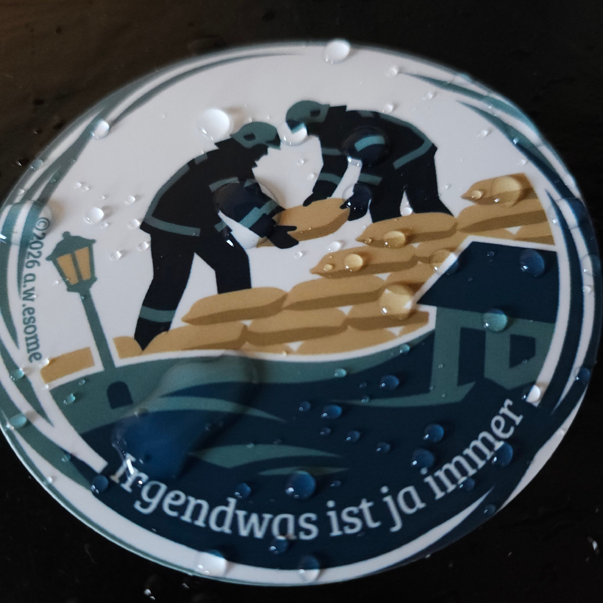Wassertropfen auf rundem Feuerwehr-Sticker zum Sammeln. Motiv zeigt Einsatzkräfte mit Sandsäcken beim Hochwasserschutz.