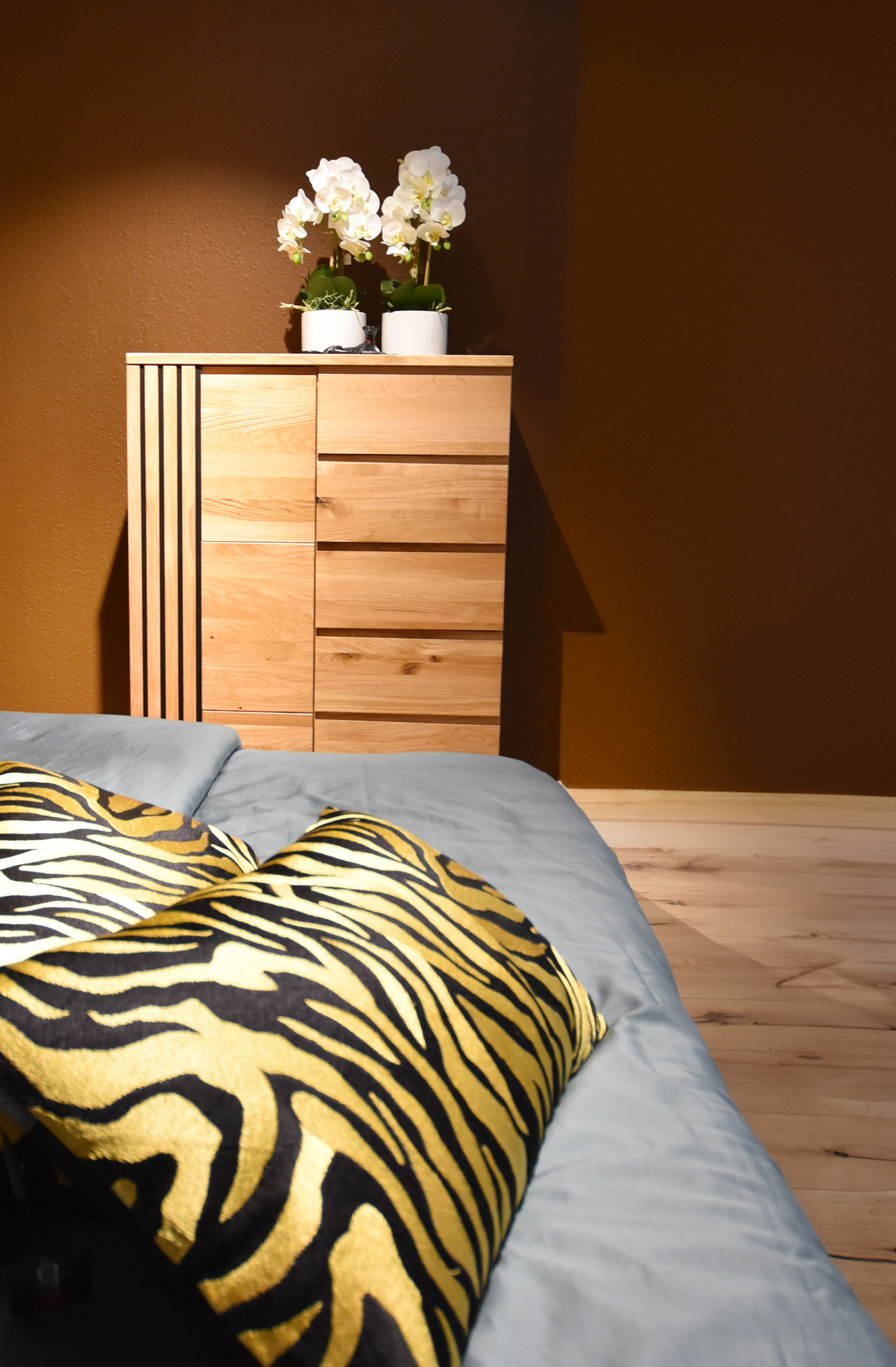 Kommode vor brauner Wand, weiß-gelbes Zebra-Kissen auf blauem Bett.