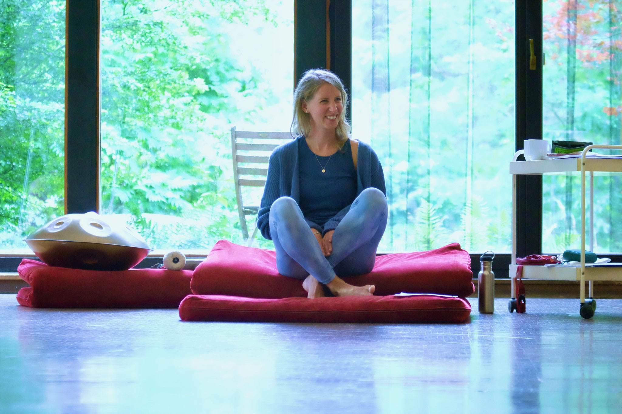 Frau (Maxine Morast) lächelnd auf Meditationskissen sitzend mit Handpan neben ihr und Ausblick ins Grüne beim Seminar zur bewussten Ernährung und Achtsamkeit in der Grube Louise im Westerwald