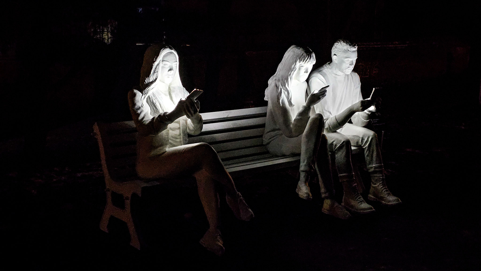 In "Absorbed by Light" von Gali May Lucas sitzen drei Menschen auf einer Bank. Ihre Köpfe sind gebeugt, ihre Finger tippen und wischen, und ihre Gesichter werden von den Bildschirmen ihrer Telefone erleuchtet.