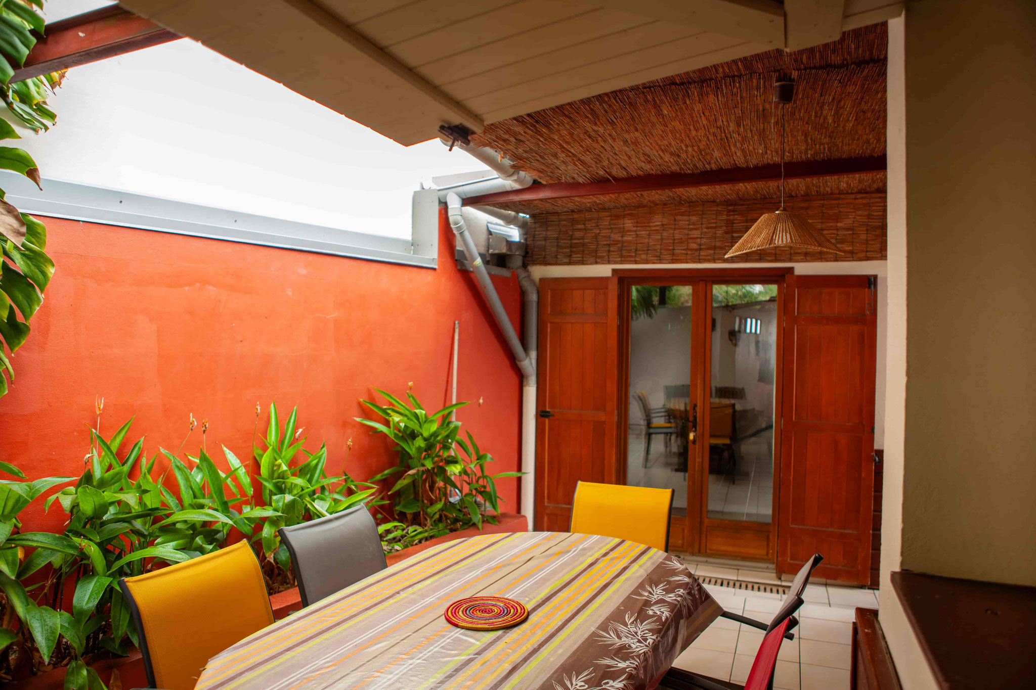 Vibrant covered outdoor dining terrace with tropical plants and orange wall, Bungalow 10 T4, Le Cardinal des Grands Bleus