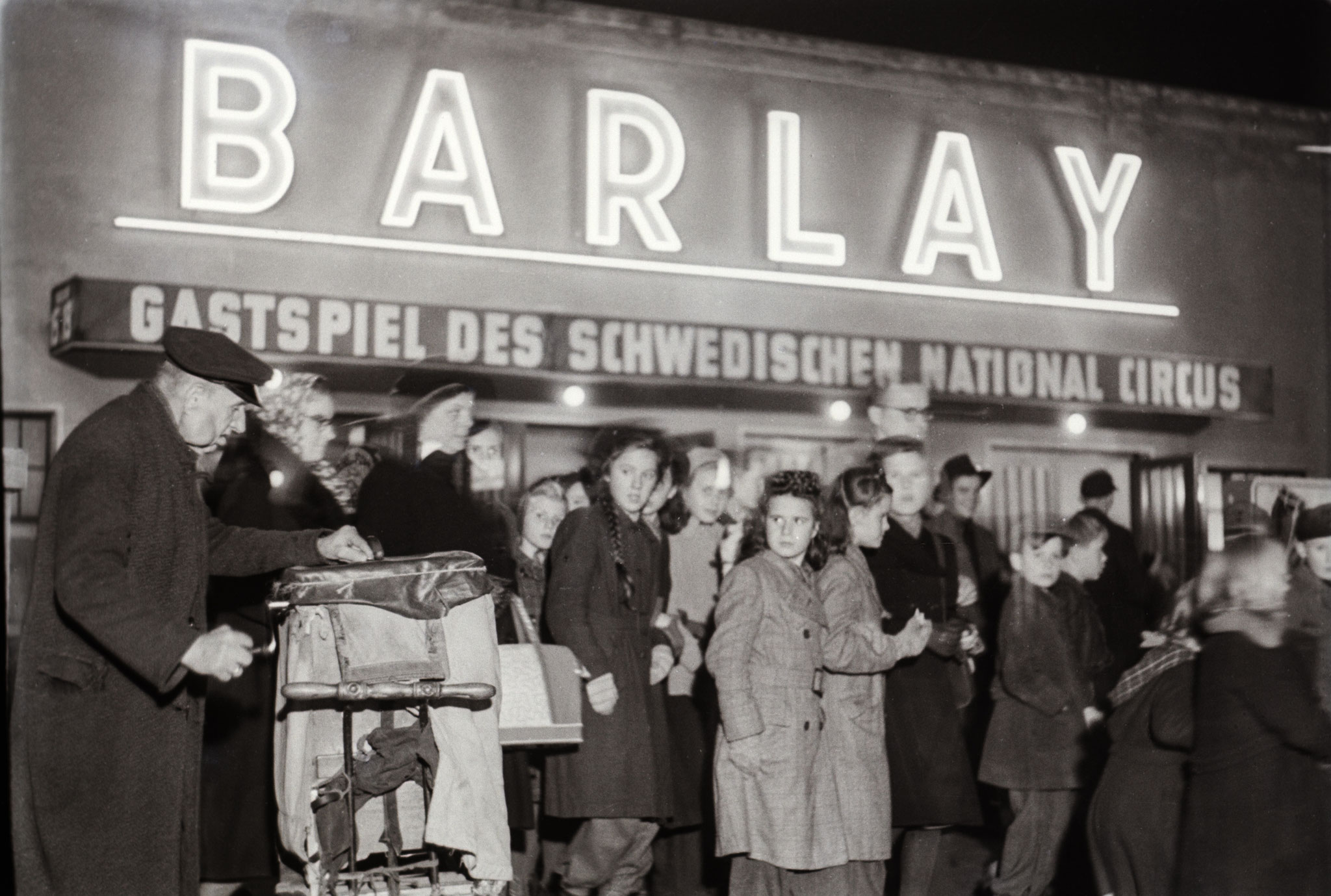 Ein Leierkastenmann steht am Eingang vor Besuchern des Zirkus Barlay in der Friedrichstrasse. Der Zirkus nahm 1947 seinen Spielbetrieb wieder auf und wurde später in den Staatszirkus der DDR eingegliedert. Foto: DDR Fotoerbe / Eva Richter