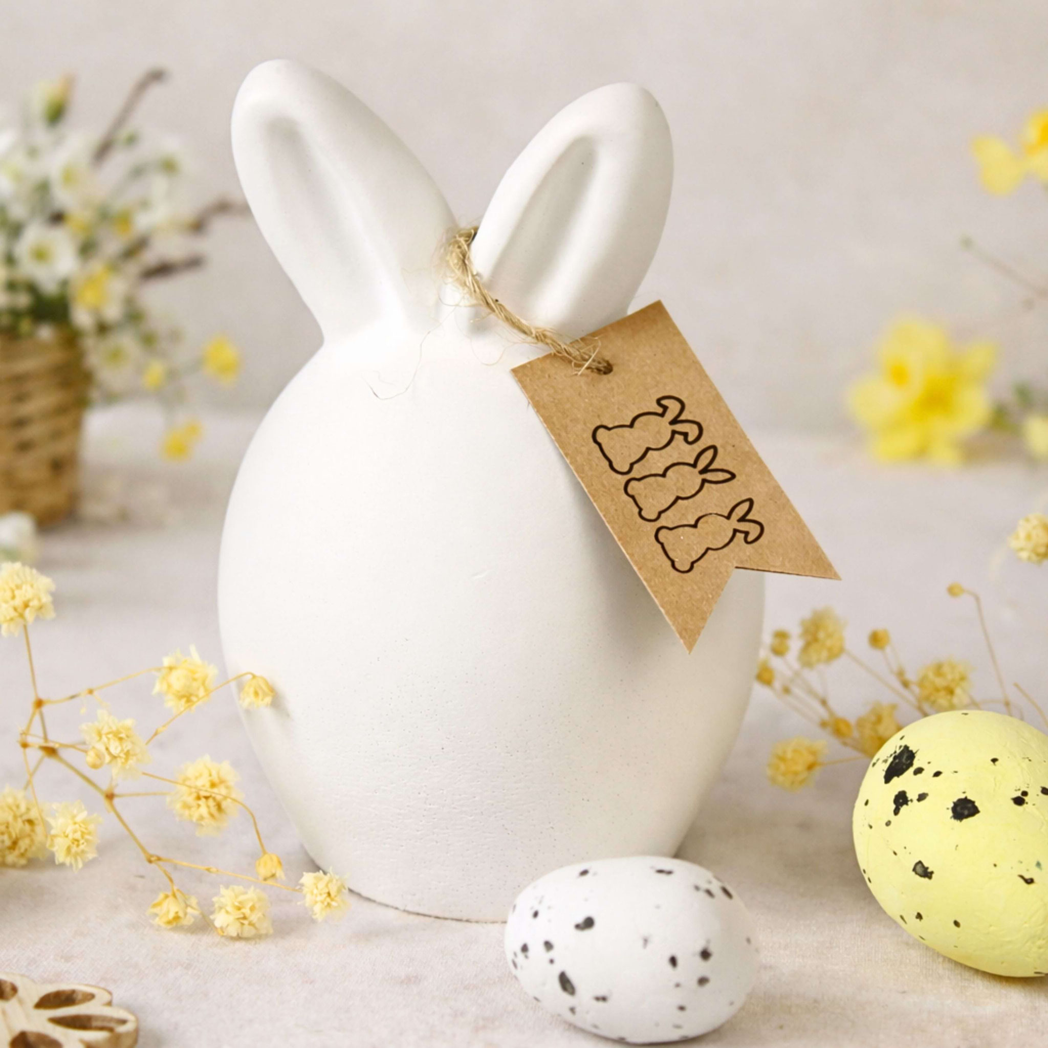 Osteranhänger aus Kraftpapier mit Hasenmotiv an Keramikhase, dekoriert mit Eiern und Frühlingsblüten.