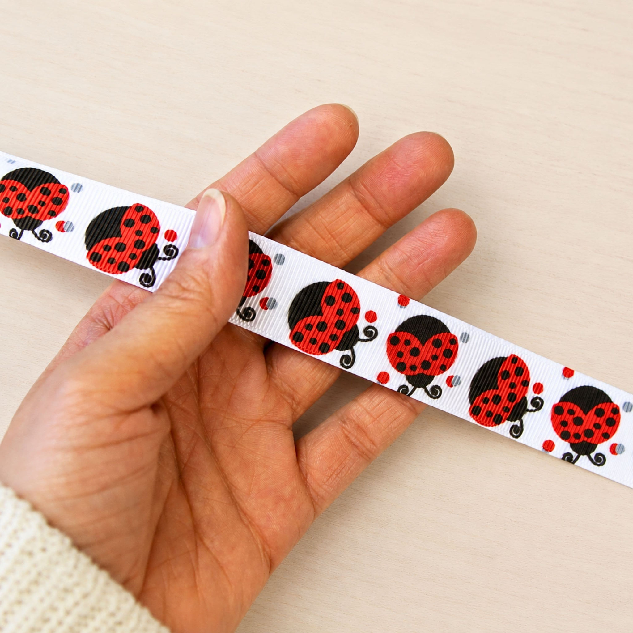 Hand hält weißes Ripsband mit Marienkäfer-Motiv zur Veranschaulichung der Breite für Näh- und Bastelprojekte.