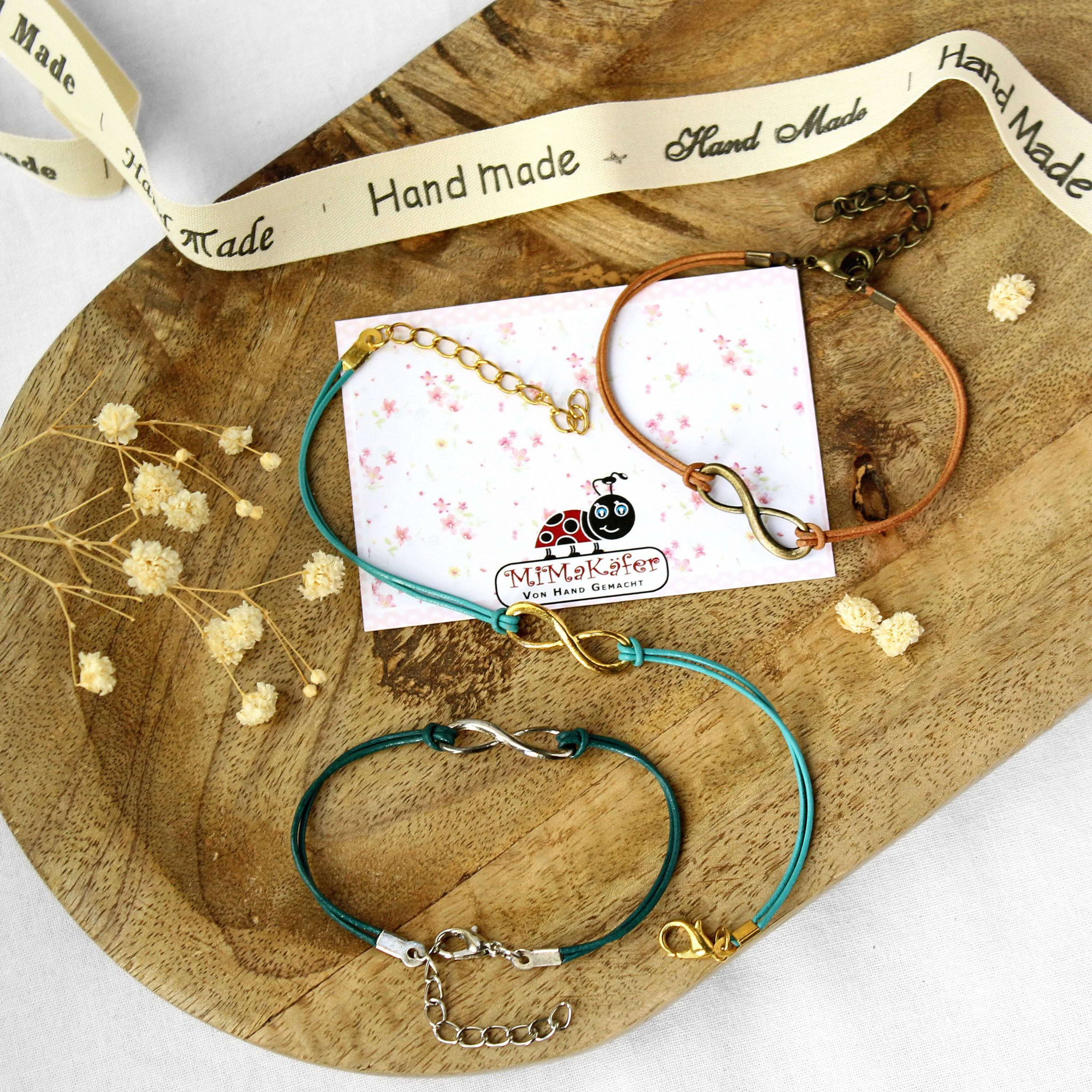 Handgemachte Infinity‑Armbänder in drei Farben auf Holztablett mit Deko, präsentiert als liebevoller Handmade‑Schmuck für Geschenke.