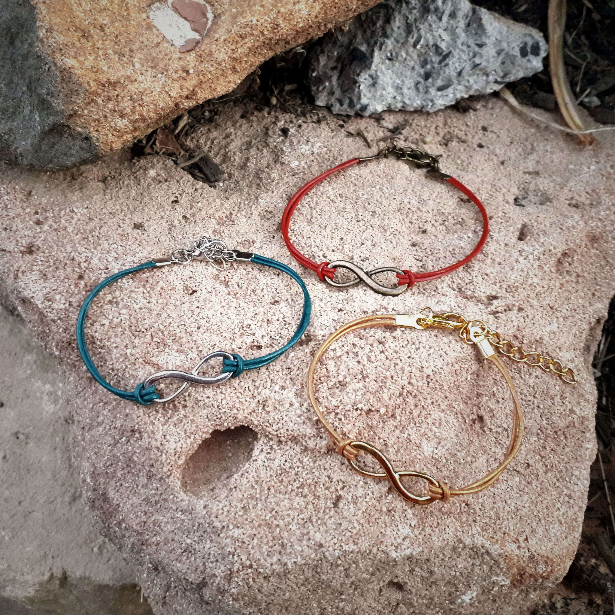 Handgemachte Infinity‑Armbänder in Rot, Türkis und Beige auf Steinoberfläche, gezeigt als moderne, personalisierbare Schmuckauswahl.