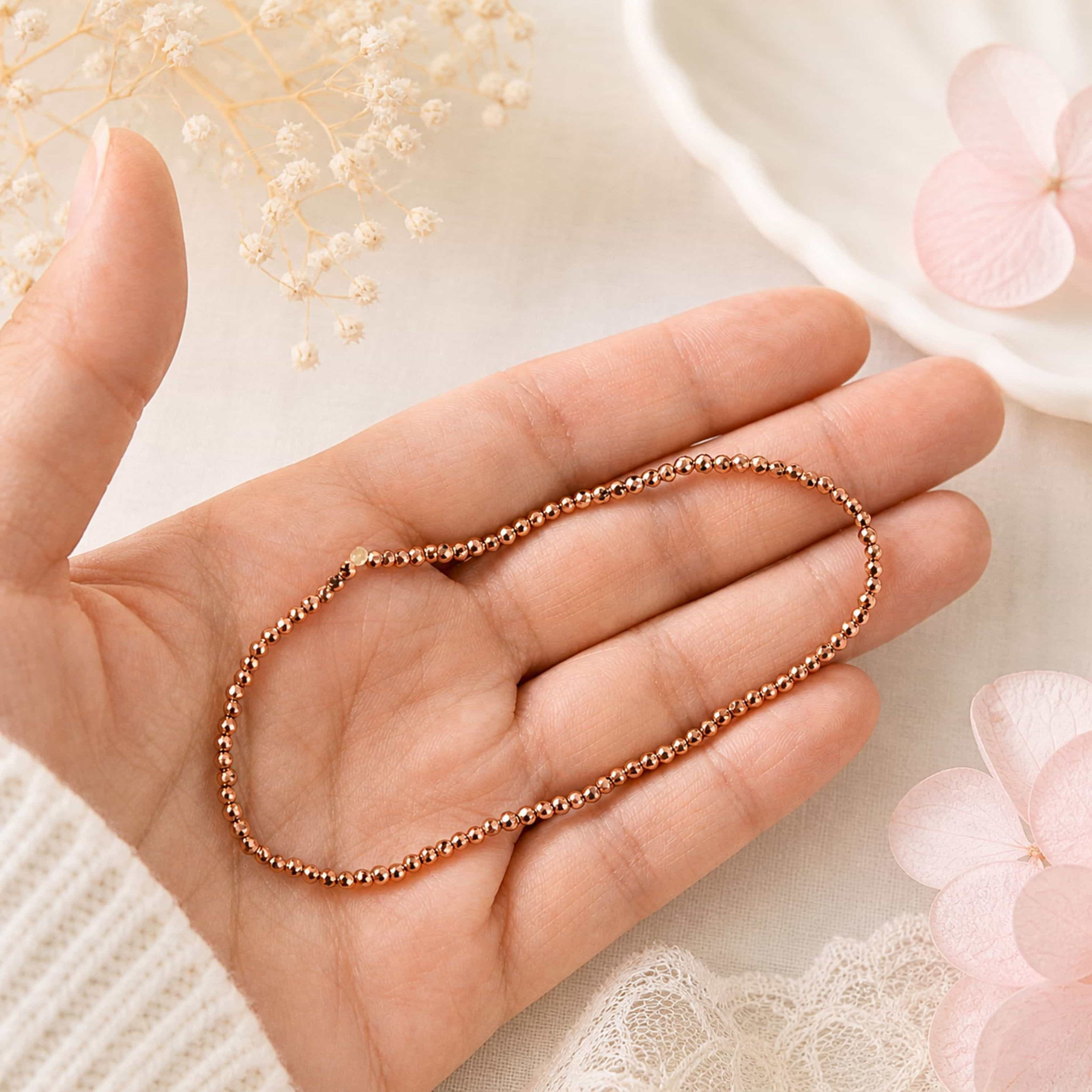 Hand hält roségoldenes Perlenarmband vor zarter Deko.