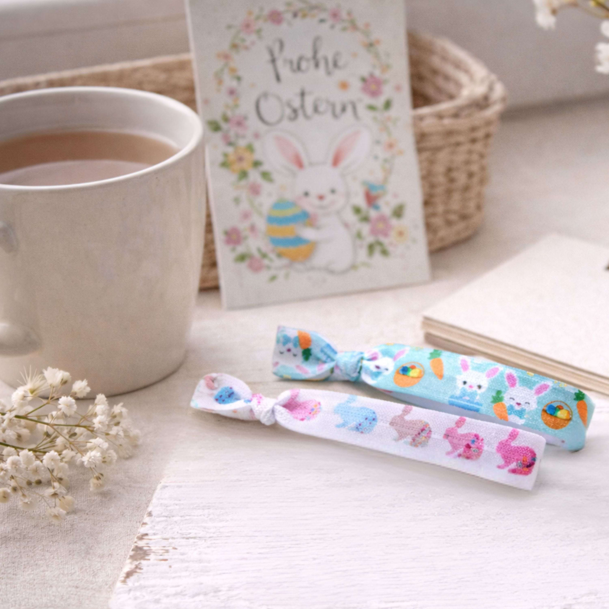 Zwei elastische Osterarmbänder mit Hasenmotiven neben Kaffeetasse und Osterkarte „Frohe Ostern“.