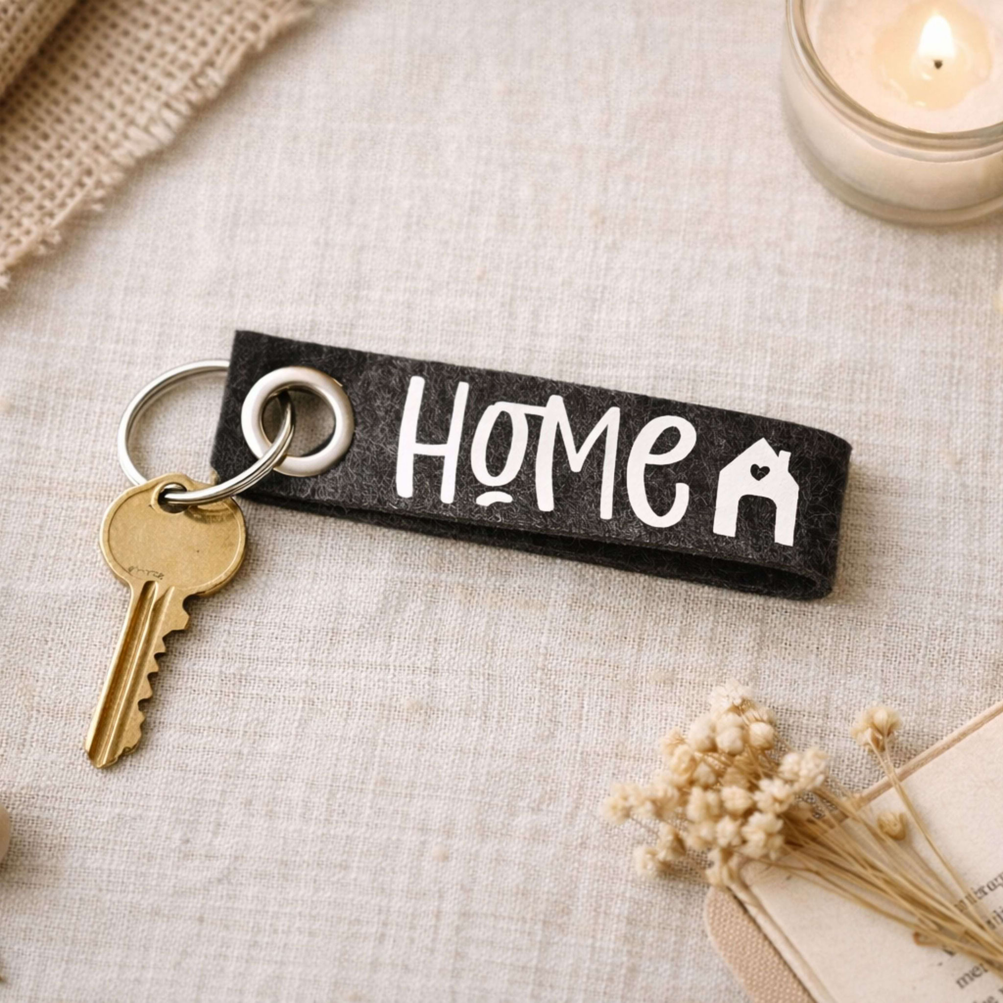 Schlüssel mit „HOME“-Anhänger auf Stoff, umgeben von Kerze, Trockenblumen und Karte – stimmungsvolles Geschenk-Arrangement.