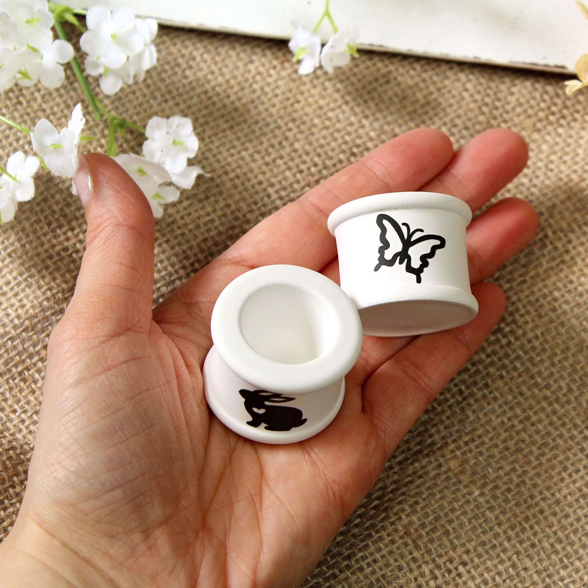 Handgehaltene weiße Kerzenhalter mit Hase- und Schmetterlingmotiv, dekorativ präsentiert als Frühlings- und Osterdeko.