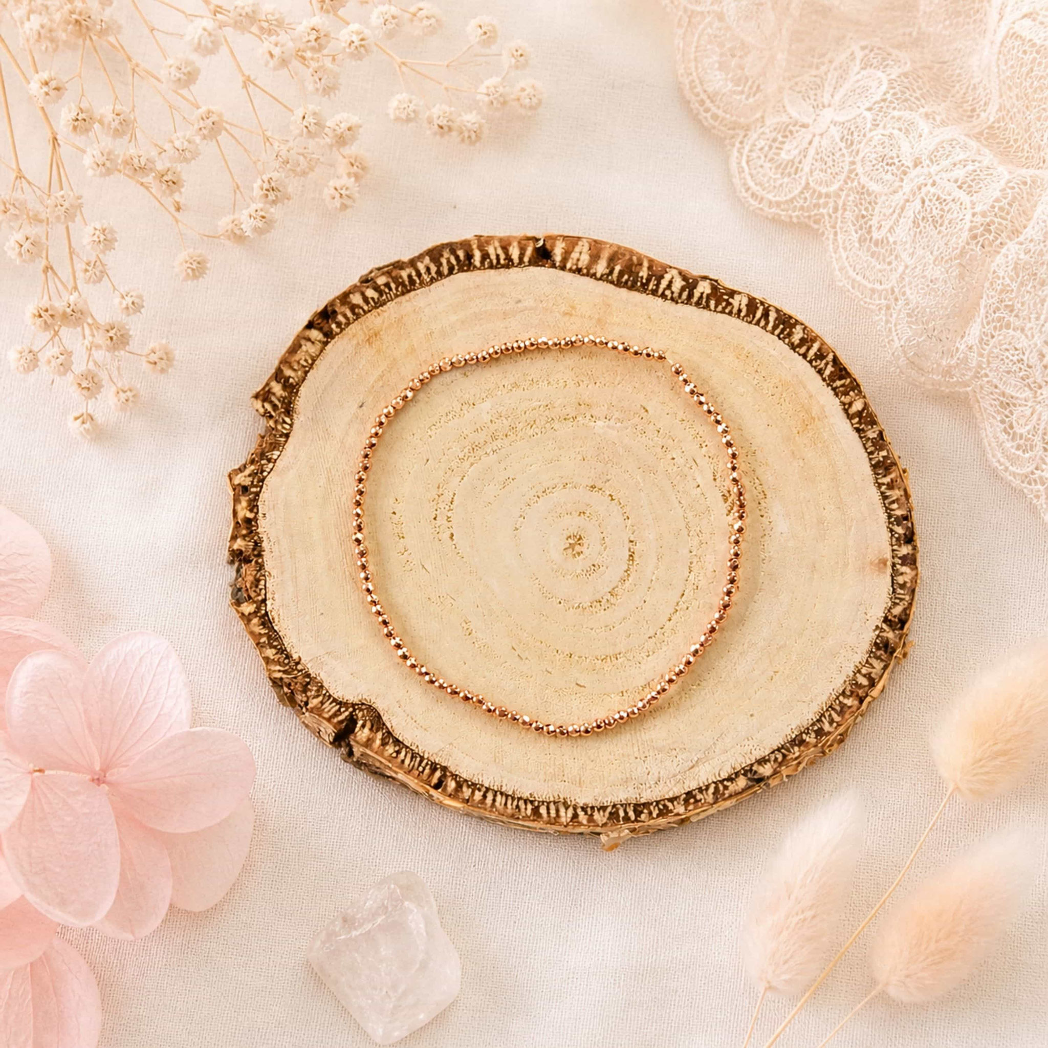 Roségold-Armband auf Holzscheibe mit Trockenblumen und Spitze.