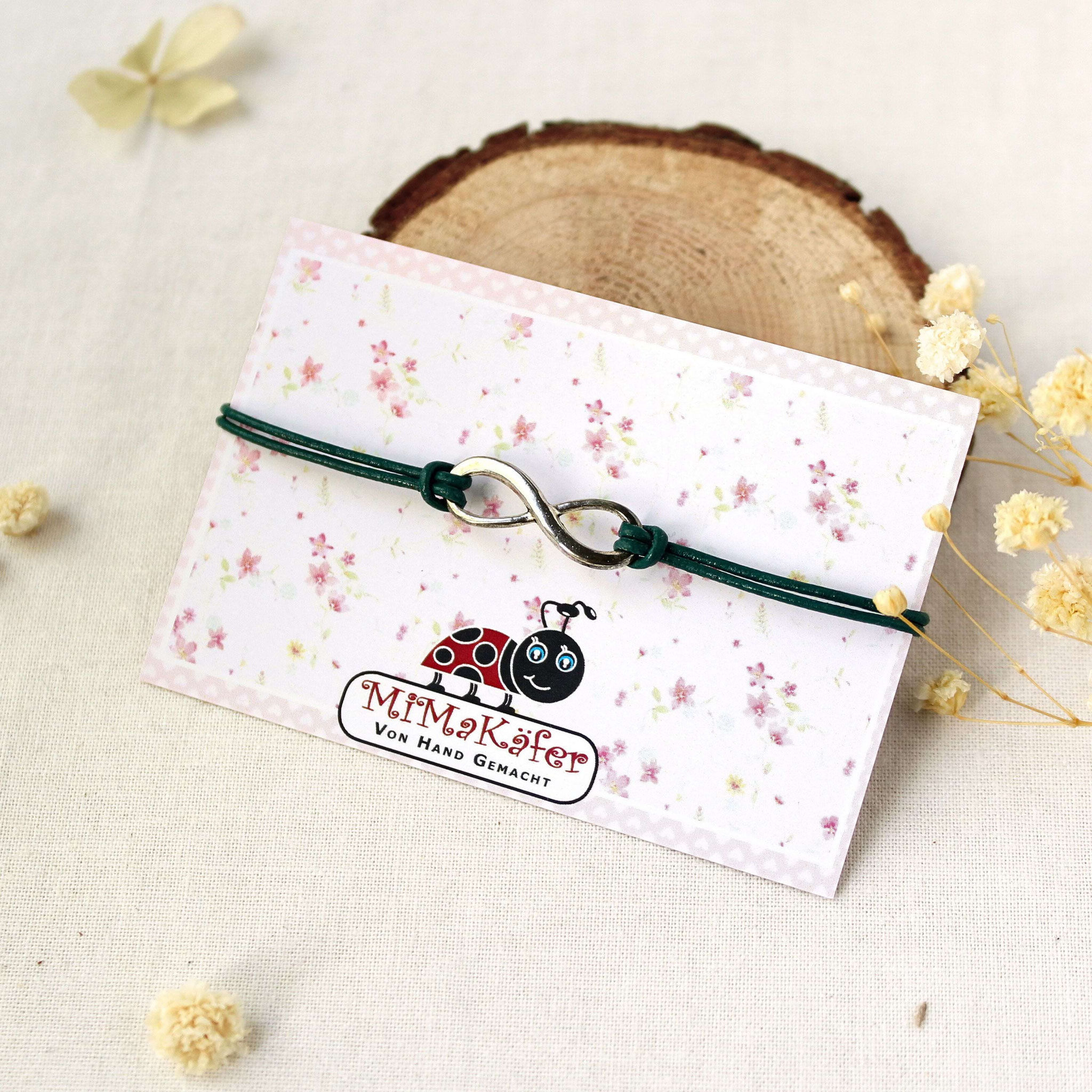 Handgemachtes Infinity‑Armband mit grünem Band auf floraler Karte, präsentiert als liebevoller, personalisierbarer Handmade‑Schmuck.