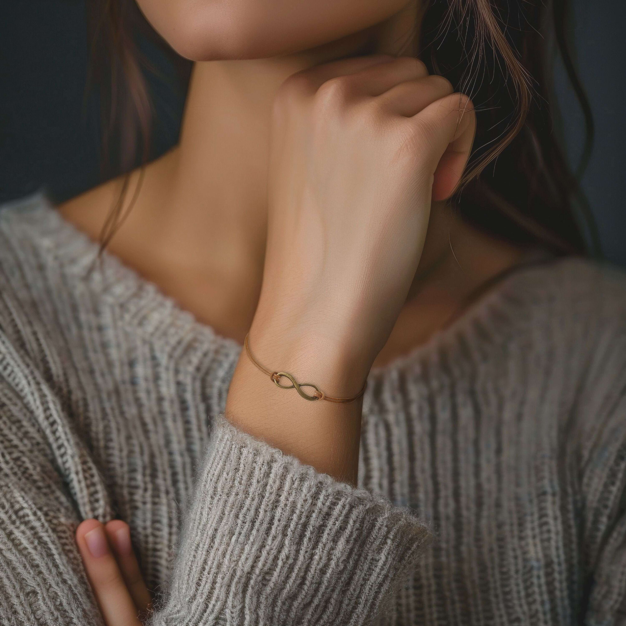 Ein zarter Fokus auf ein goldfarbenes Infinity-Armband am Handgelenk, getragen zu einem grauen Strickpullover als Schmuck‑Lifestyle‑Detail.
