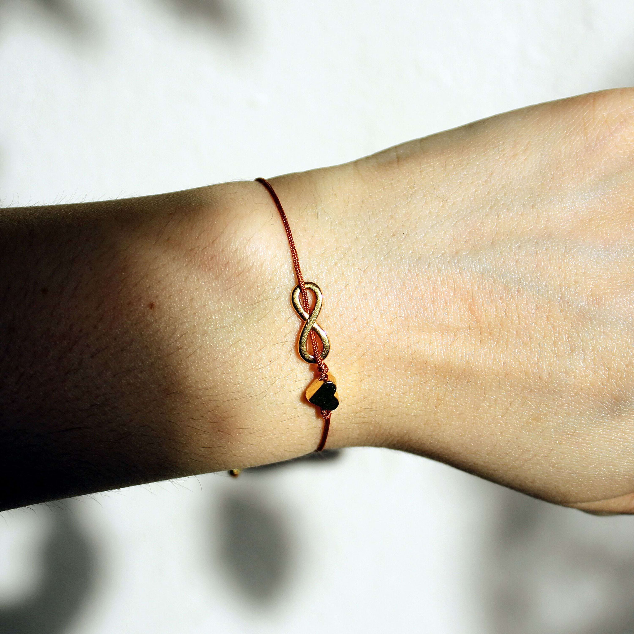 Nahaufnahme eines roten Makramee‑Armbands mit goldener Infinity‑ und Herzplakette, getragen am Handgelenk.