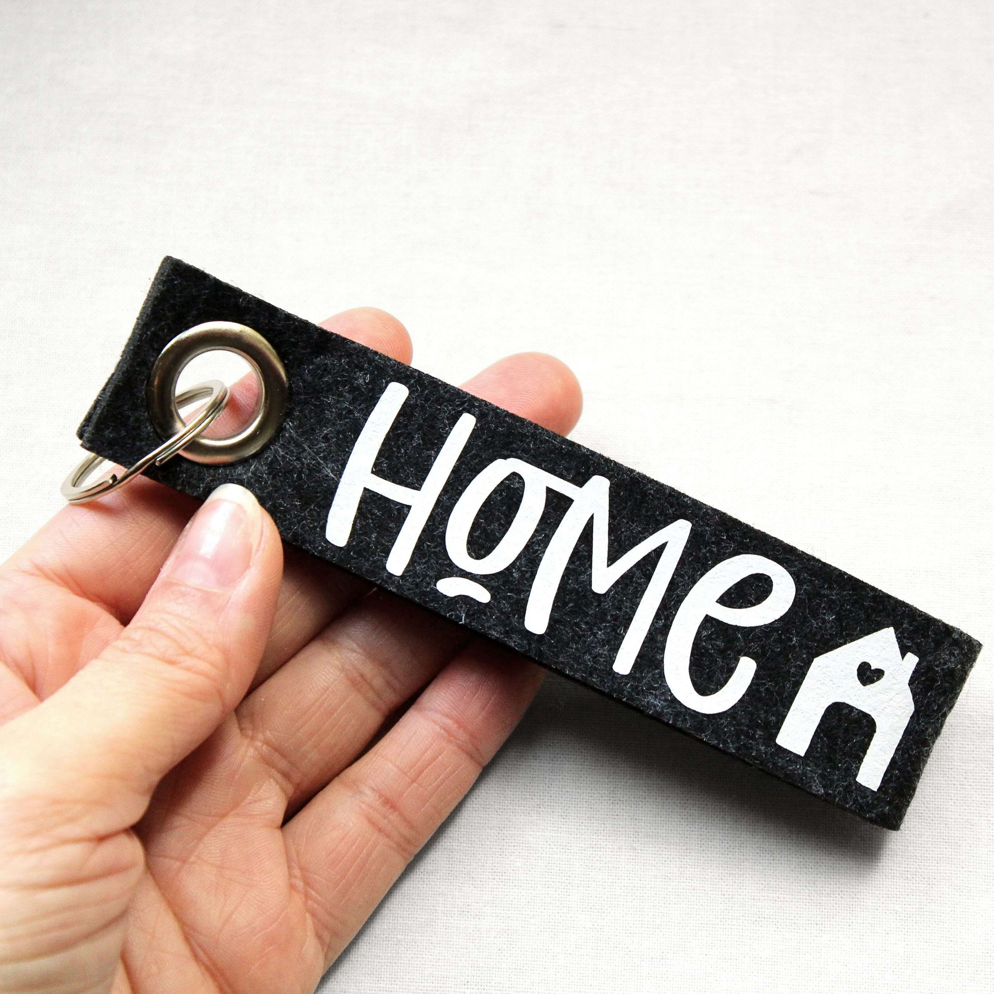 Hand hält schwarzen Filz-Schlüsselanhänger mit „HOME“-Print und Herzhaus-Symbol – liebevolles Accessoire für Zuhause.