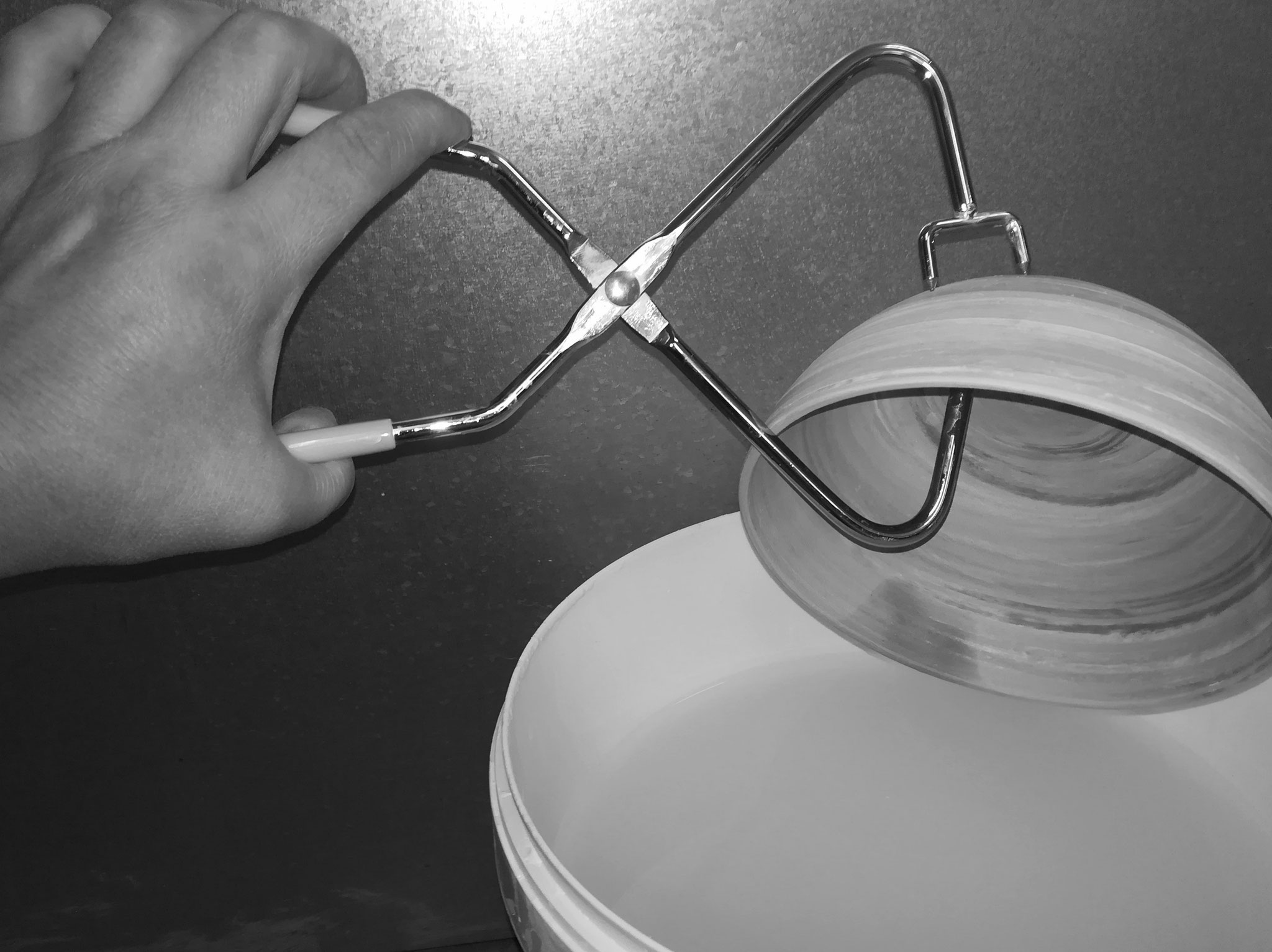 eine Hand hält mit einer Glasierzange eine Schüssel über einen Eimer mit Glasur