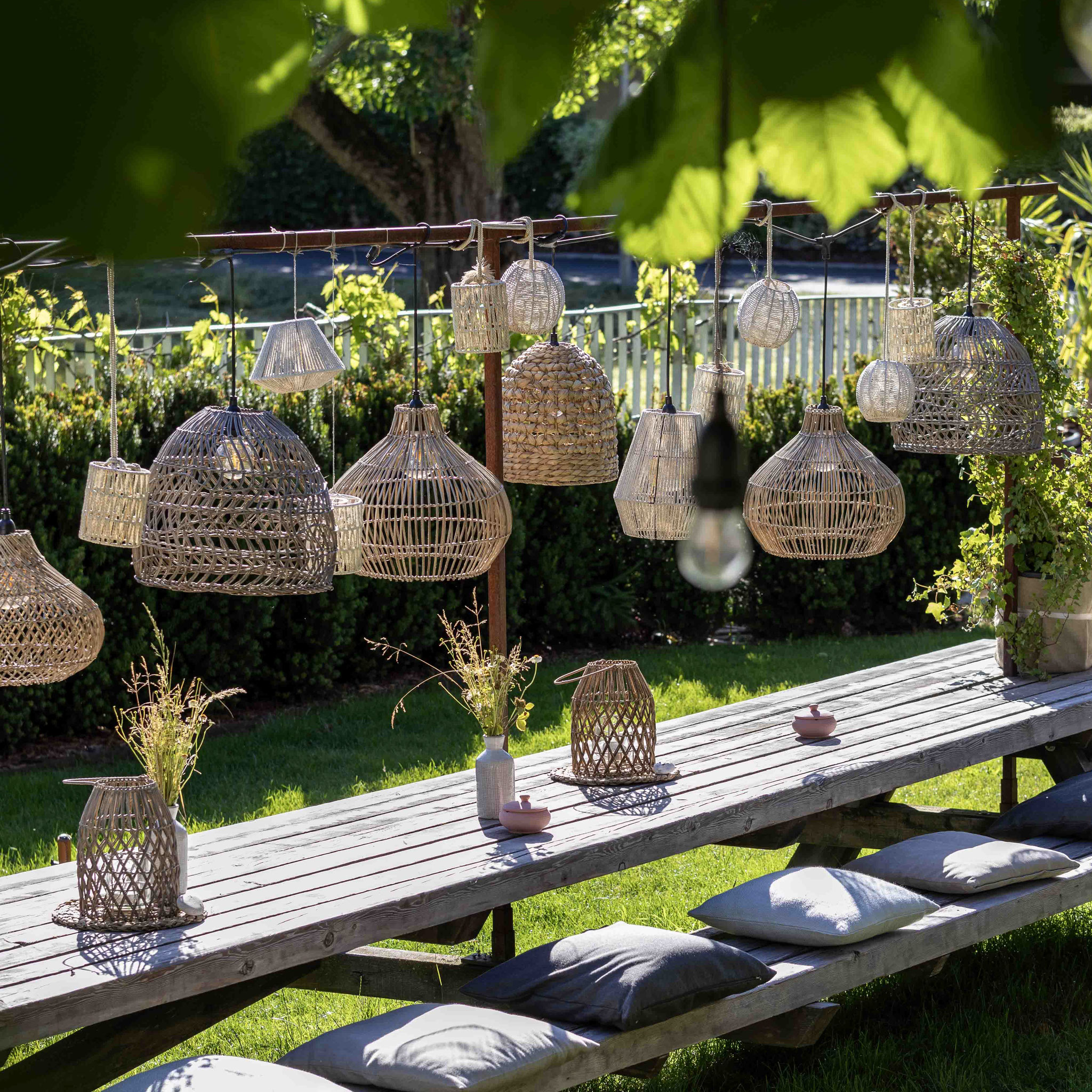 Rustikale Holztafel mit Sitzkissen unter Bäumen, dekoriert mit geflochtenen Lampenschirmen und Blumenarrangements im Garten des Boutiquehotel PETRUS in Reischach-Bruneck • Südtirol.