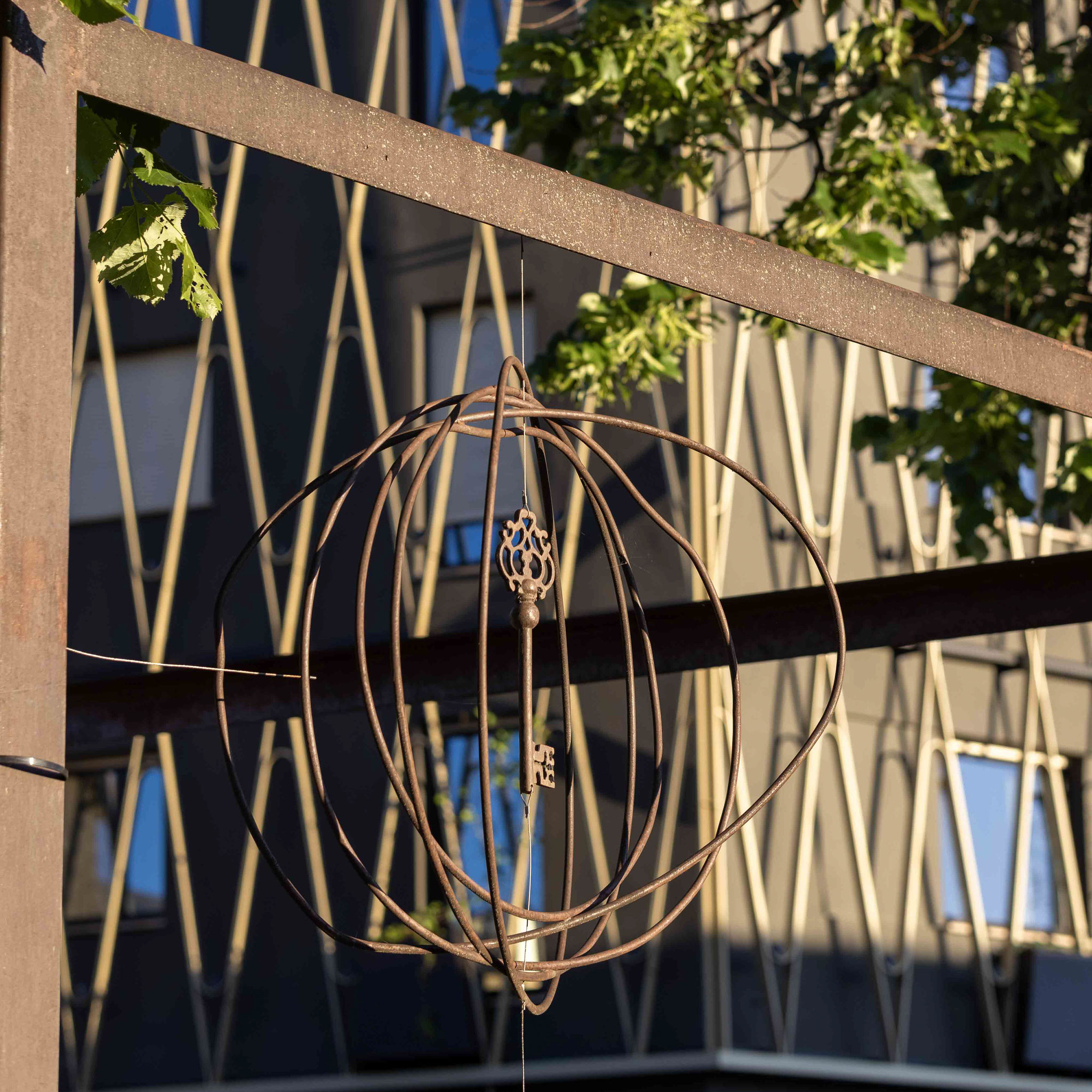 Dekoratives Metallornament in Kugelform mit einem antiken Schlüssel hängt im Außenbereich des Boutiquehotels PETRUS in Reischach-Bruneck. Im Hintergrund sind die moderne Hotelfassade mit begrünten Rankelementen und warmes Abendlicht zu sehen.
