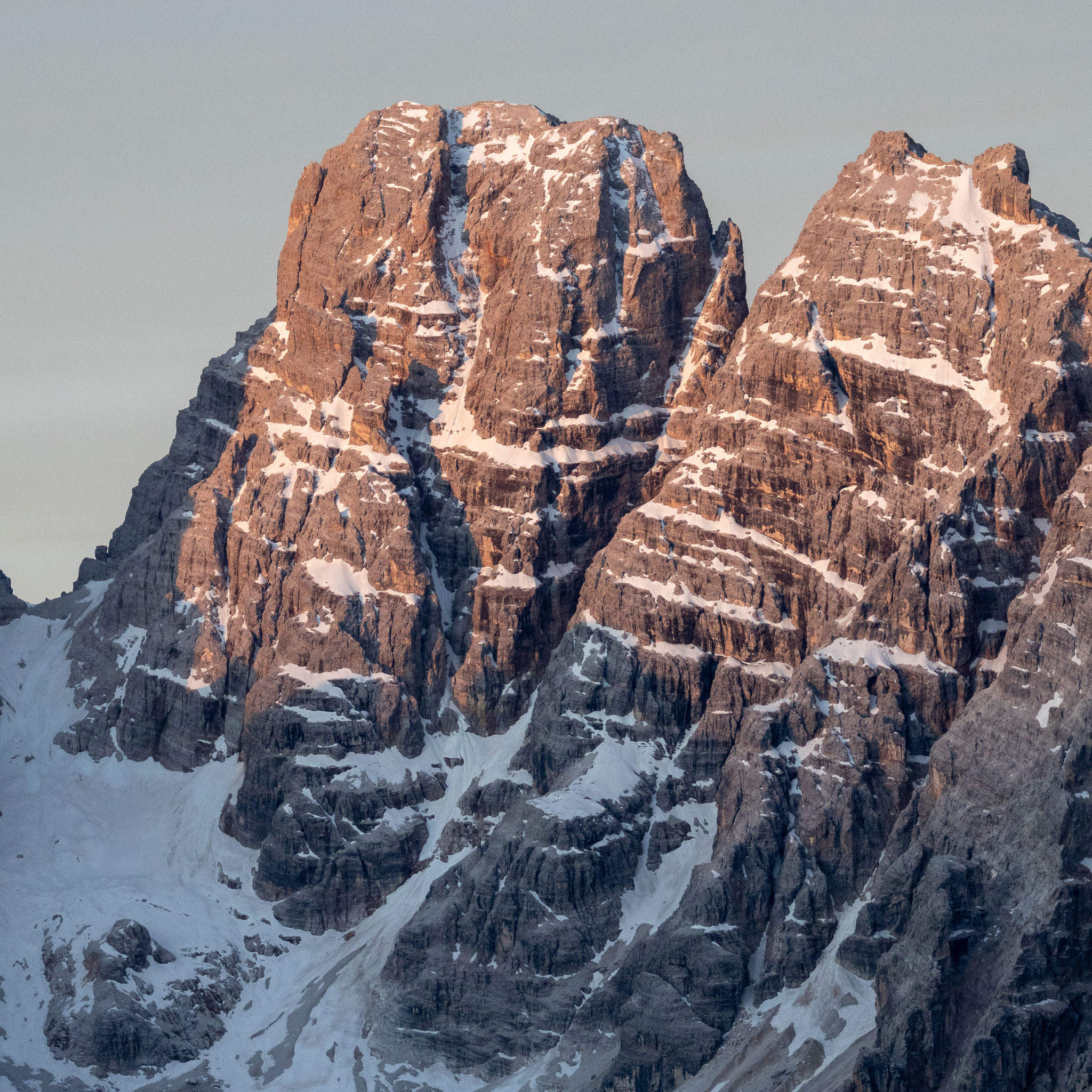 Nahaufnahme einer markanten Dolomitengruppe im warmen Licht der aufgehenden oder untergehenden Sonne. Die schroffen Felswände sind von Schneefeldern durchzogen und heben sich vor einem ruhigen, wolkenlosen Himmel in sanften Grautönen ab.